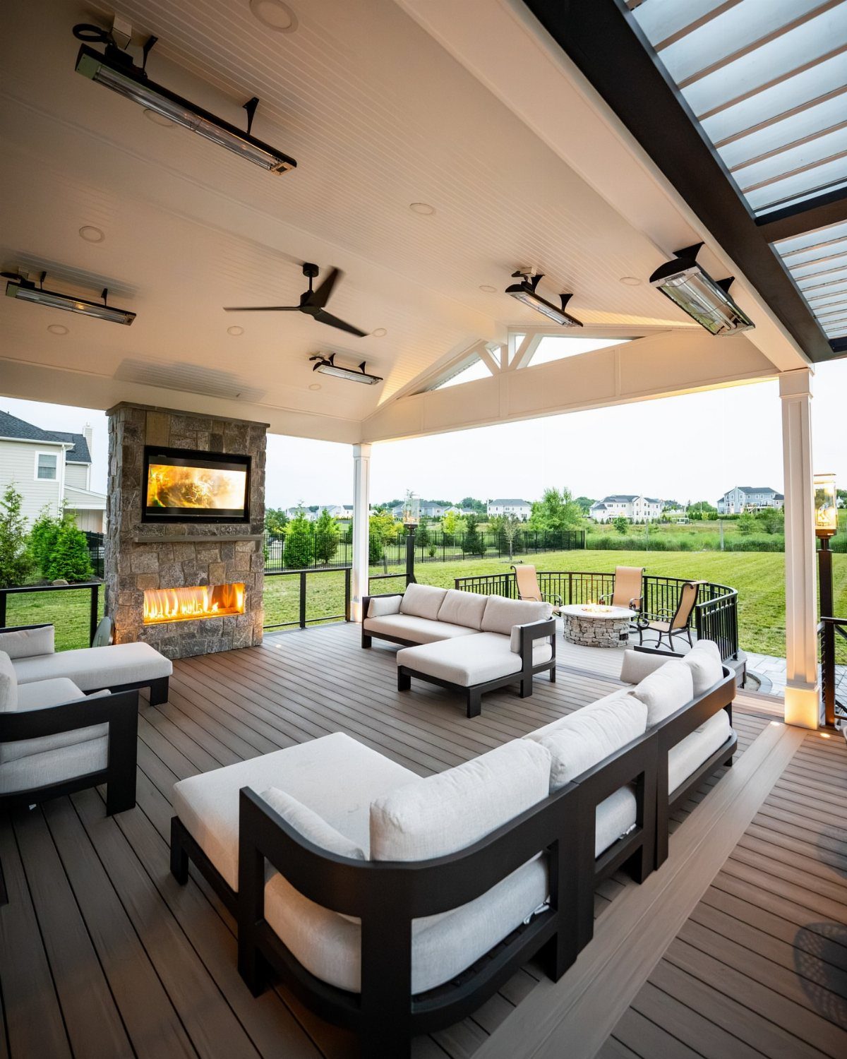 17 Backyard Deck Designs for Every Yard and Budget 17 A covered wooden deck featuring a stone outdoor fireplace and white lounge furniture overlooking a lawn.