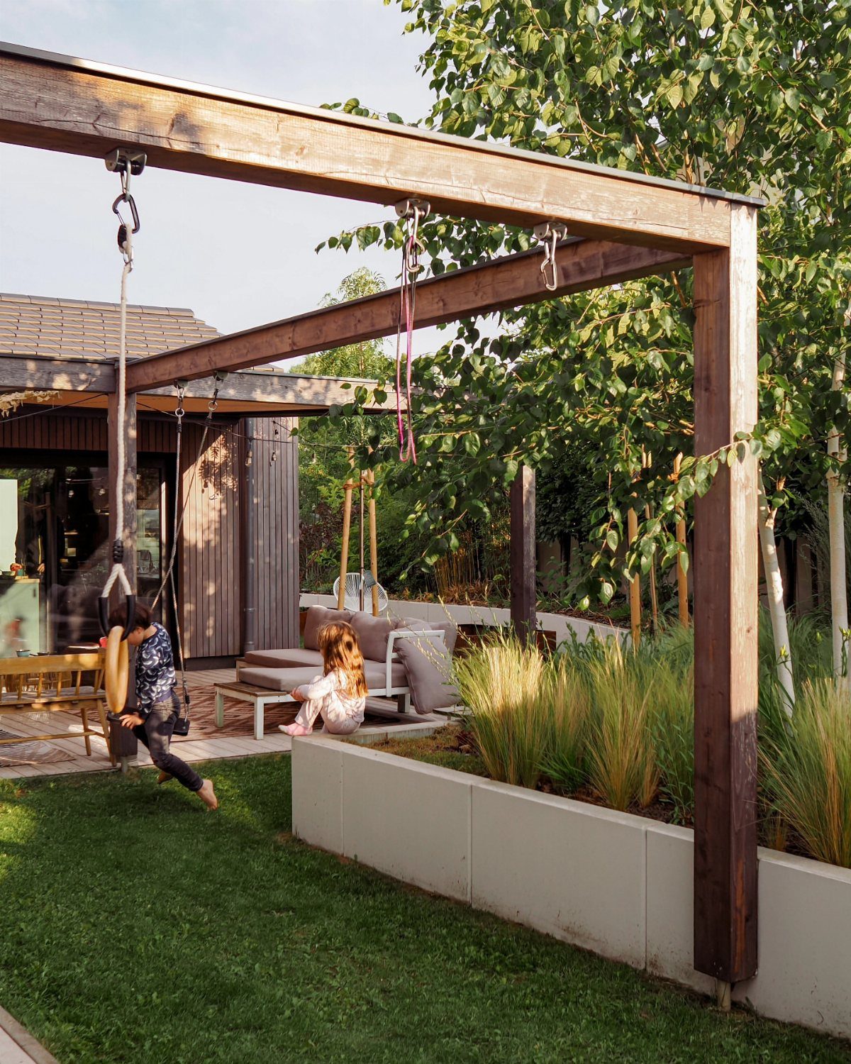 17 Backyard Deck Designs for Every Yard and Budget 13 A child sits on a swing hanging from a large wooden pergola in a backyard.