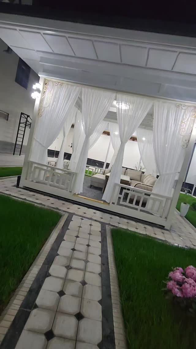 White curtained gazebo with lounge seating situated in a landscaped backyard with a stone path.