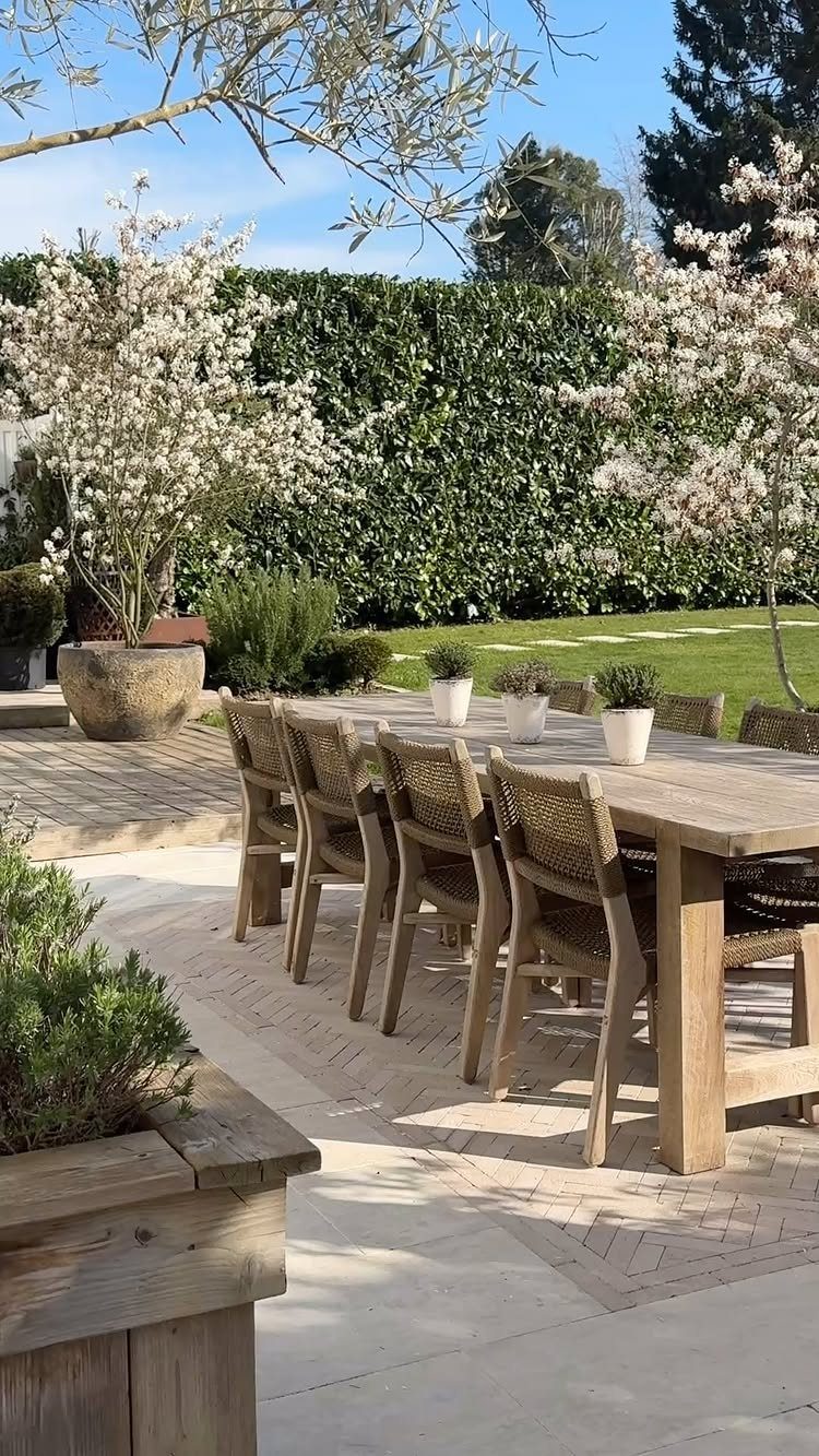 Wooden dining table and chairs set on a stone patio surrounded by greenery.