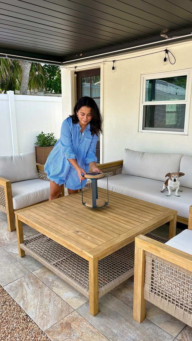 A woman in a blue dress interacts with a wooden coffee table on a furnished patio.