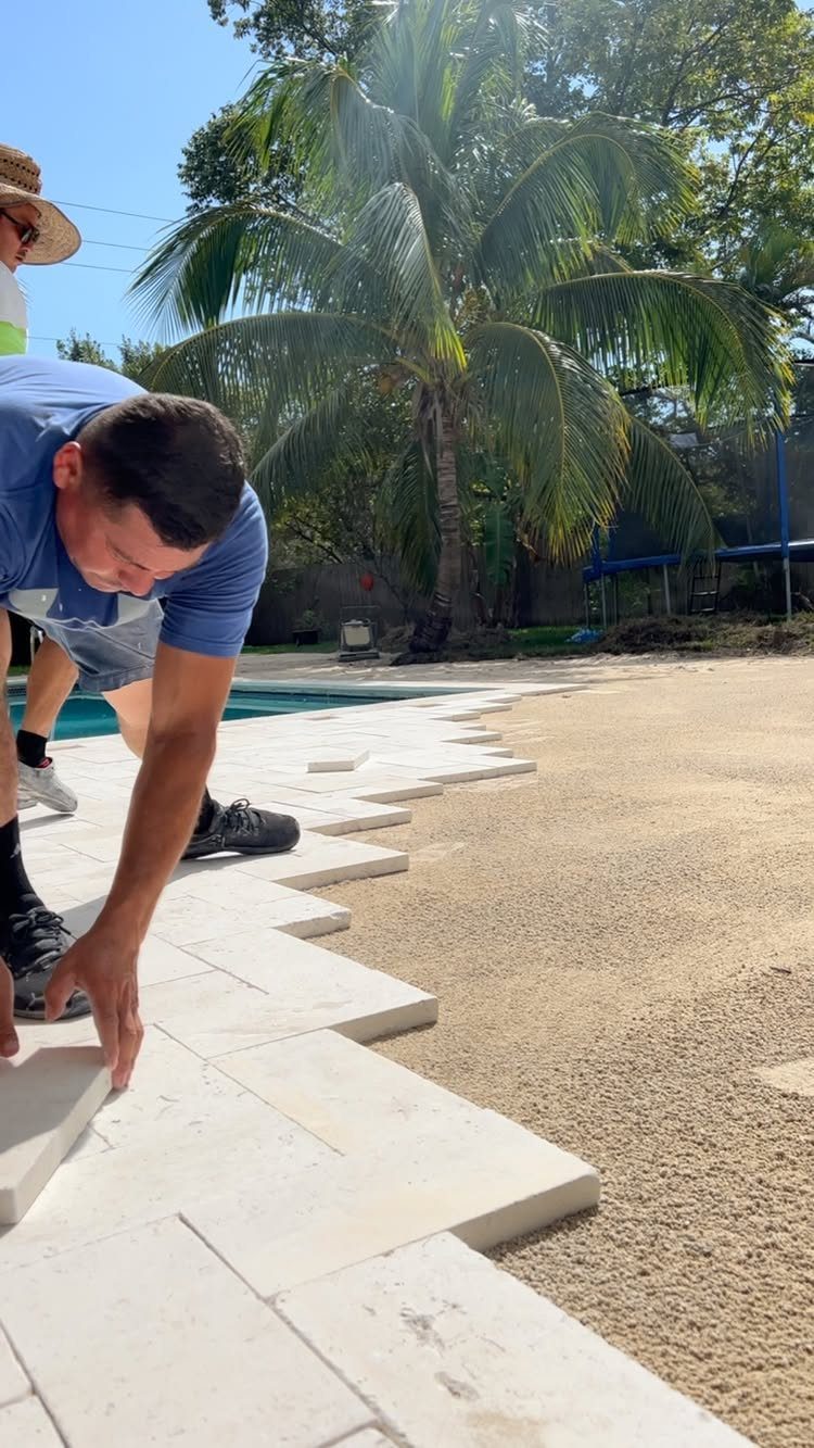 A man installs light-colored stone pavers on a sandy area next to a swimming pool.