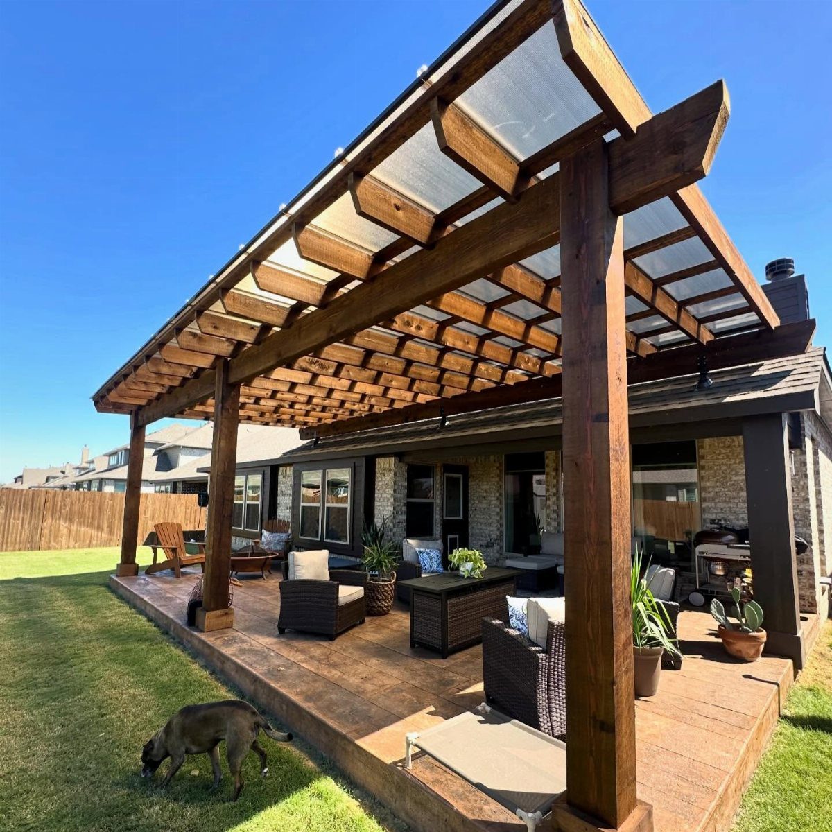 A large wooden pergola with a translucent roof provides shade over a furnished backyard patio.