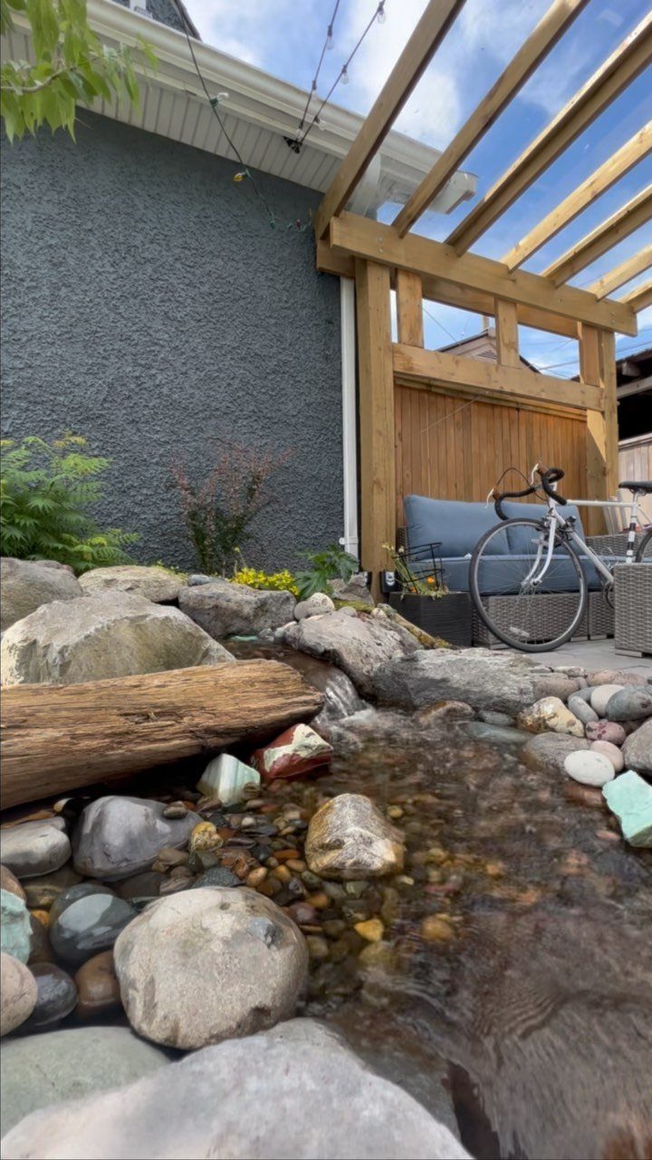 A small backyard patio with a wooden pergola, seating, and a rocky water feature.