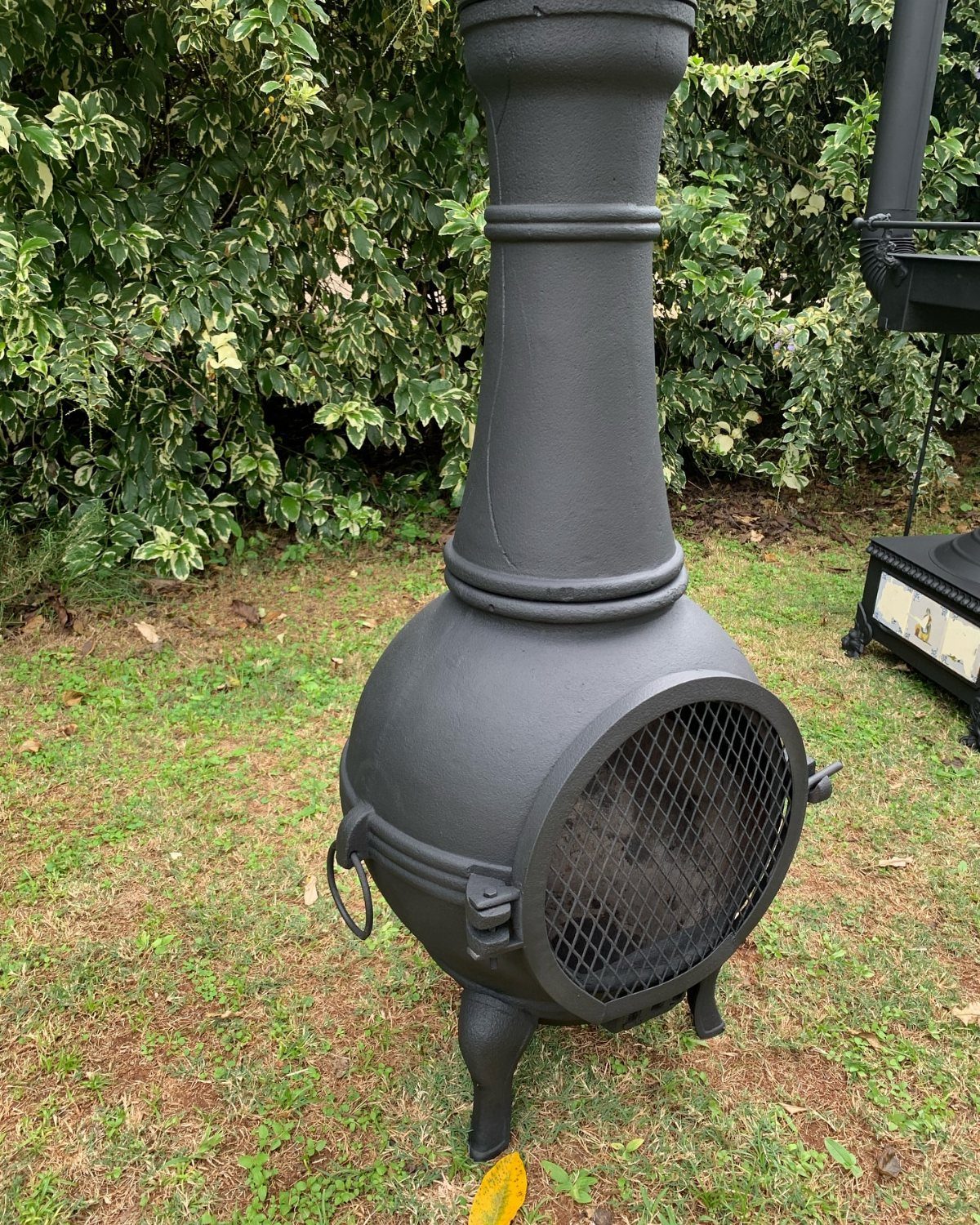 A black cast iron chiminea stands on a green grass lawn near leafy bushes.