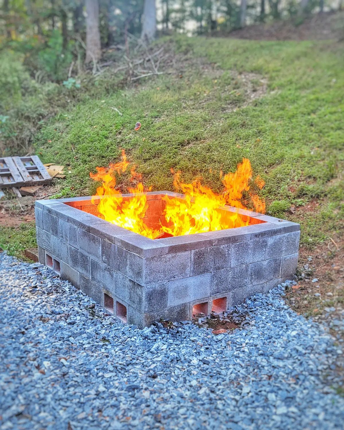 A square cinder block fire pit with bright orange flames burning inside on gravel.