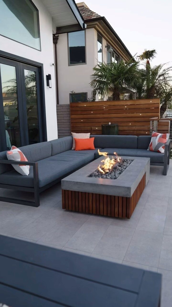 A rectangular concrete fire pit table with a wooden base sits in front of a grey outdoor sectional sofa.