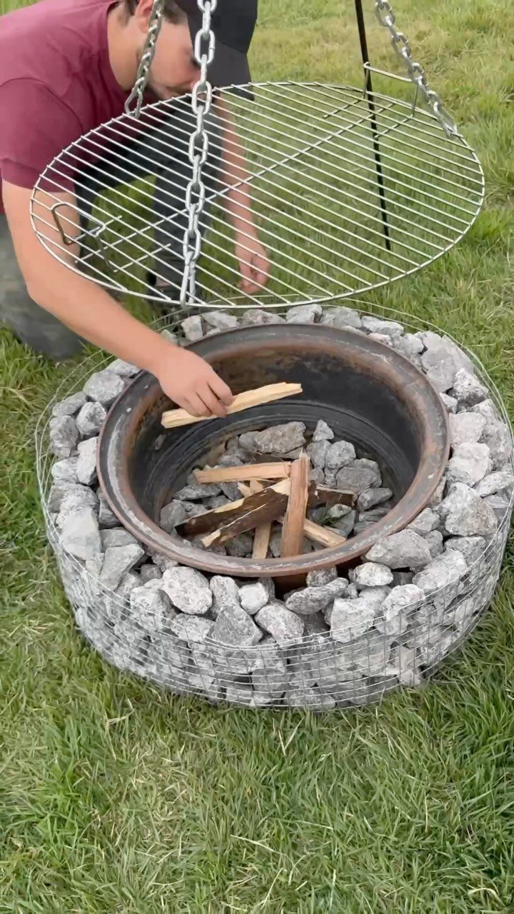 A person placing wood into a stone-filled gabion fire pit outdoors.