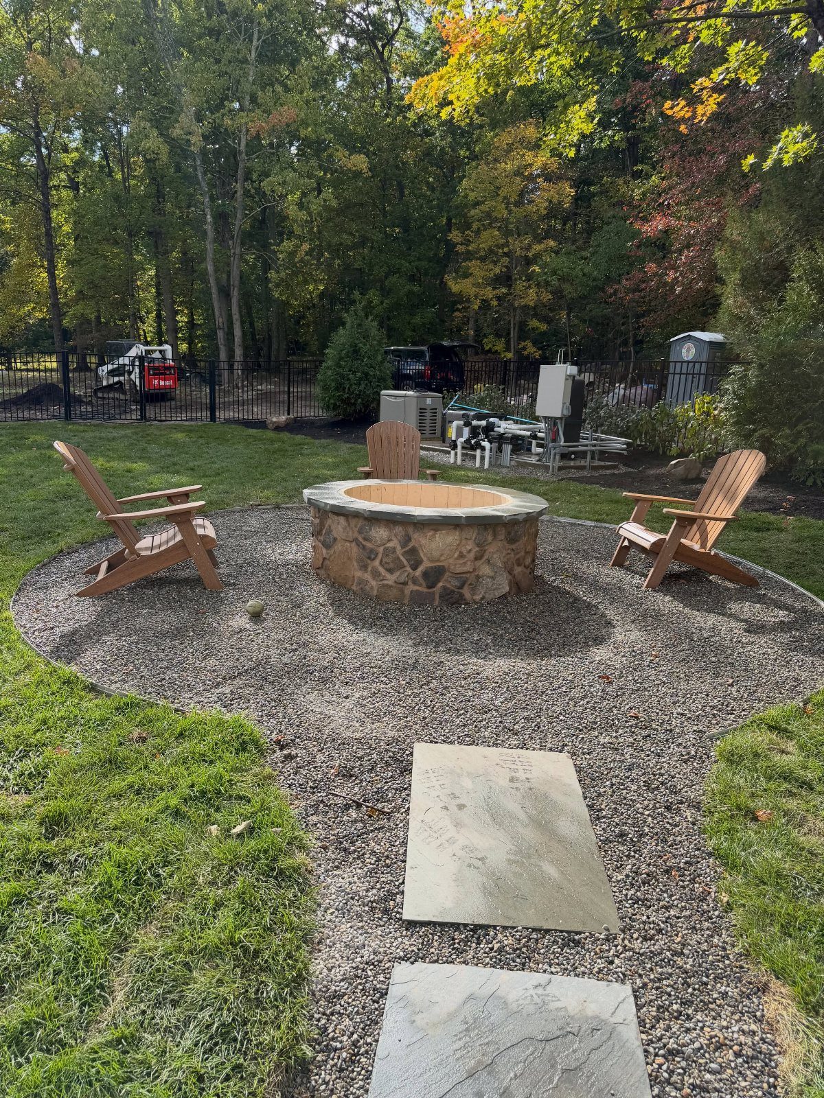A stone fire pit centered within a circular gravel area with wooden chairs and trees.
