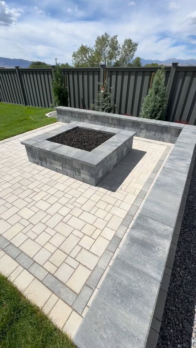 A square grey stone fire pit centered on a light-colored paver patio outdoors.