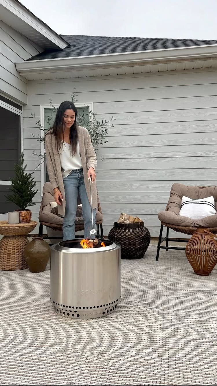 A woman tending a stainless steel portable fire pit on a patio with outdoor furniture.
