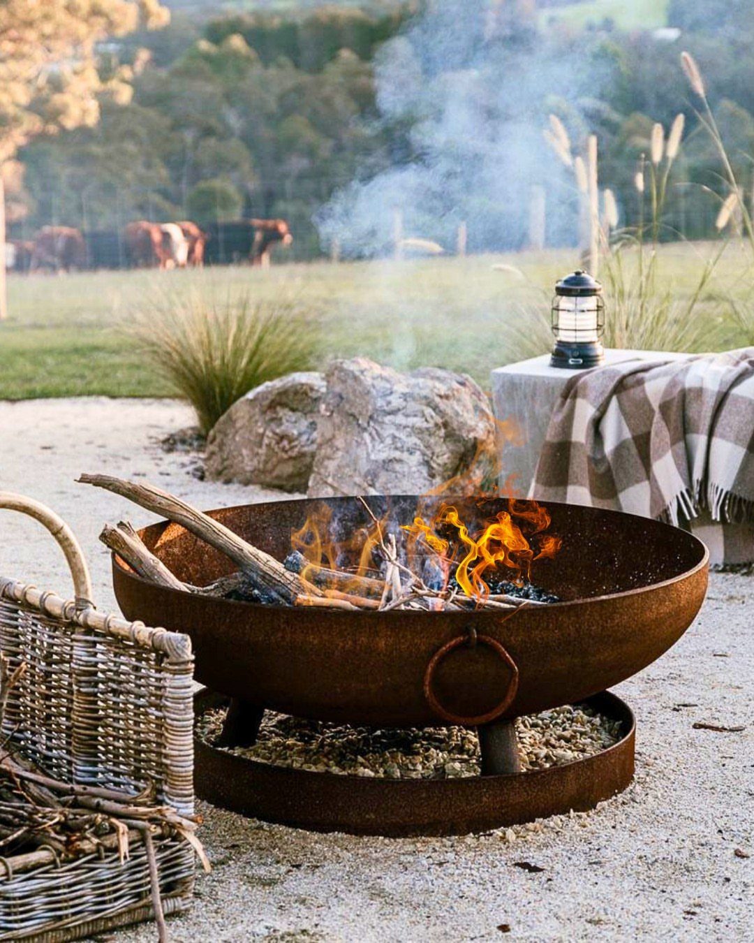 A rusty metal fire pit with burning wood logs and bright orange flames on gravel.