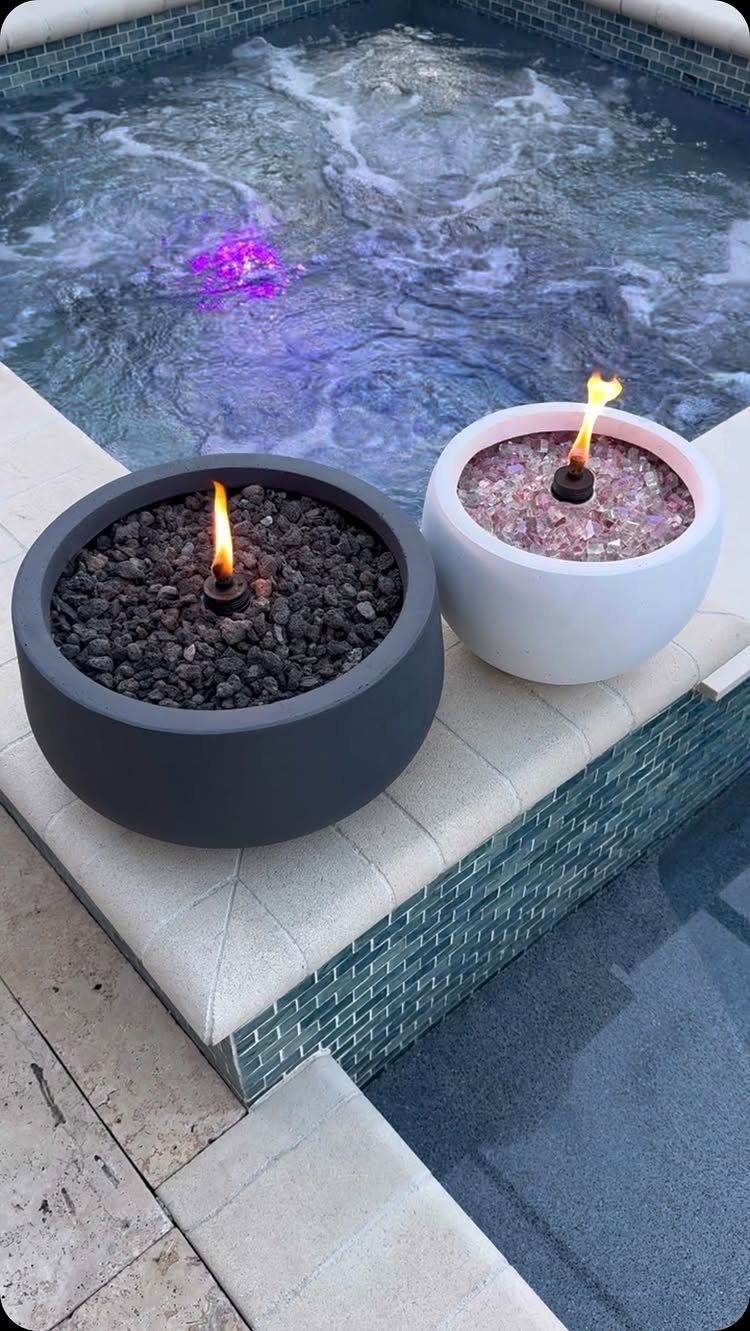 Two small tabletop fire bowls with flames sitting on the edge of a tiled swimming pool.