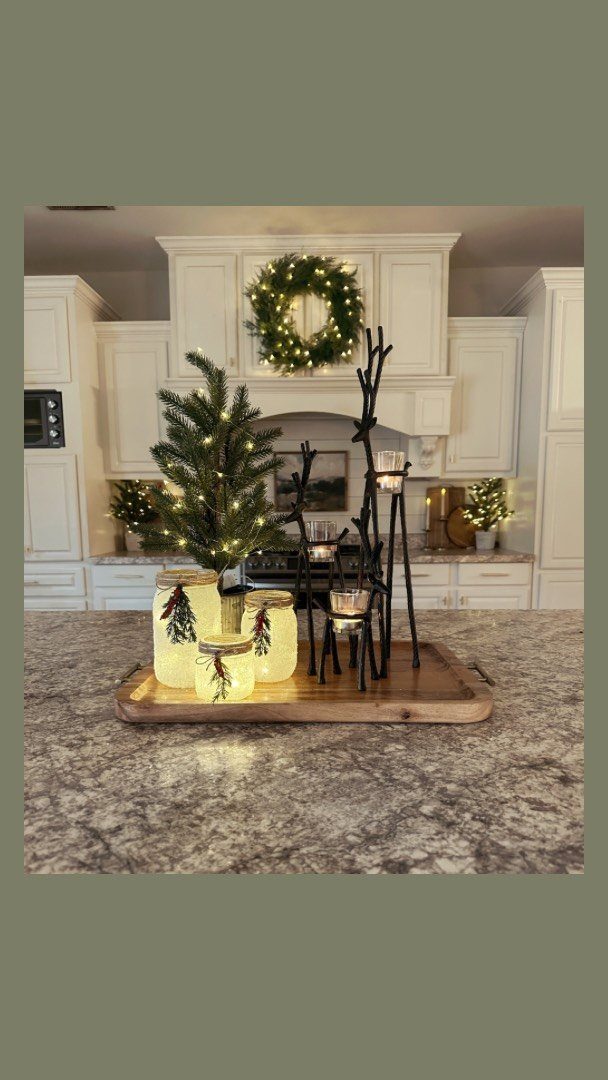 Glowing mason jars decorated with pine branches sit on a wooden tray in a kitchen.