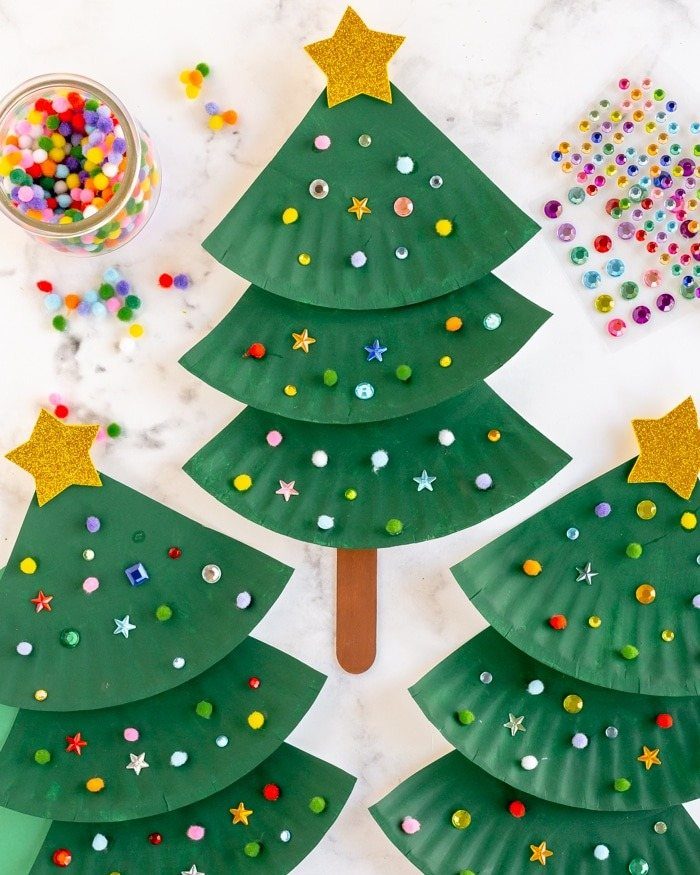 Three green paper plate Christmas trees decorated with colorful pom-poms and gold stars on marble.