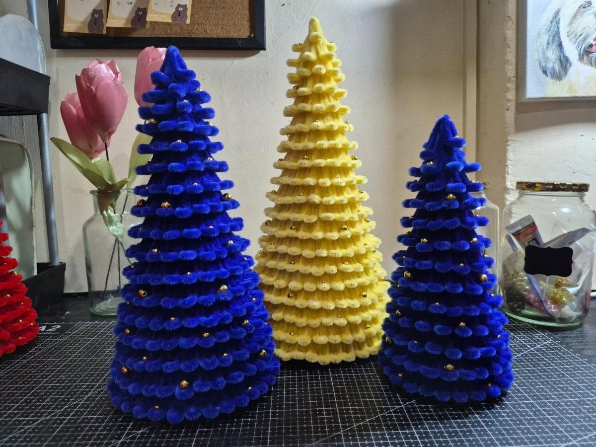 Three colorful pom-pom Christmas trees in blue and yellow standing on a dark surface.