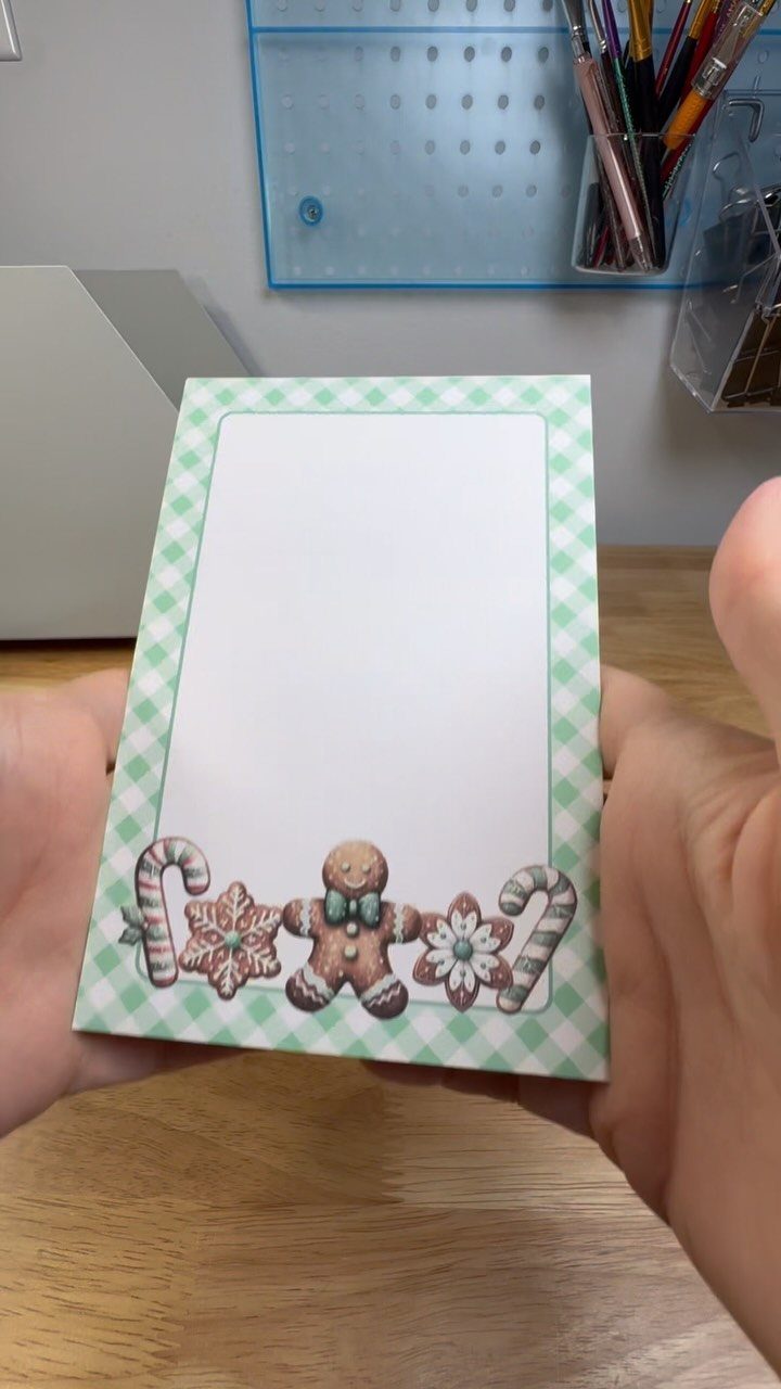Hands holding a small handmade notepad with a green checkered border and gingerbread man illustrations.