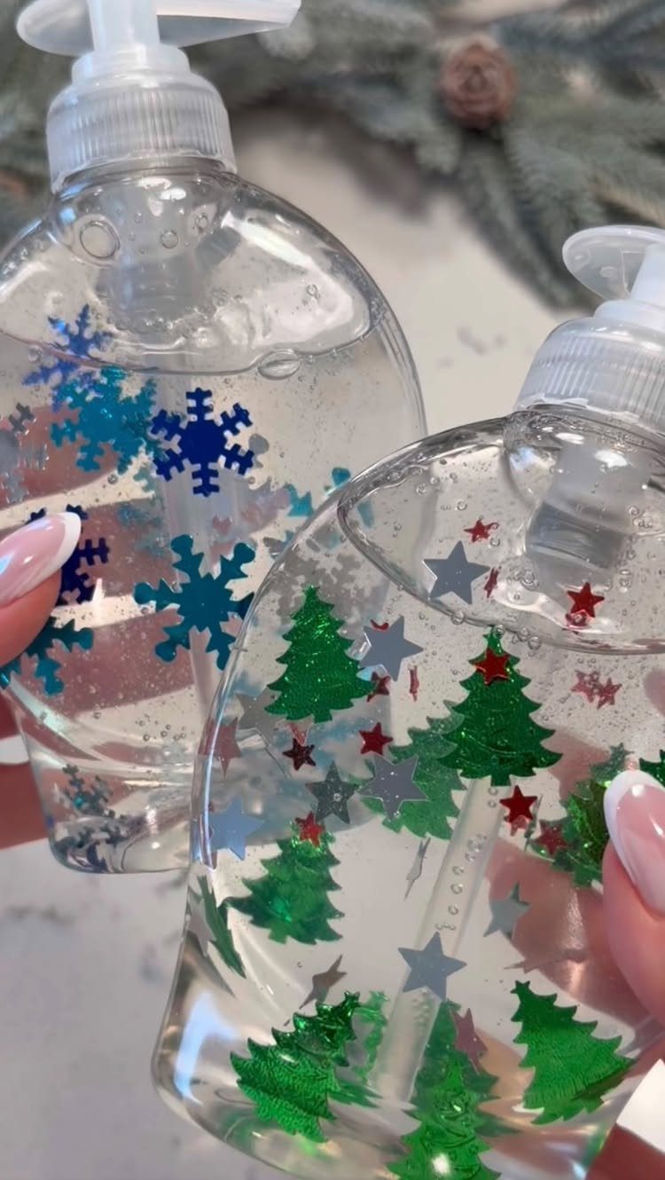 Two clear liquid soap dispensers decorated with blue snowflakes and green Christmas trees held by hands.