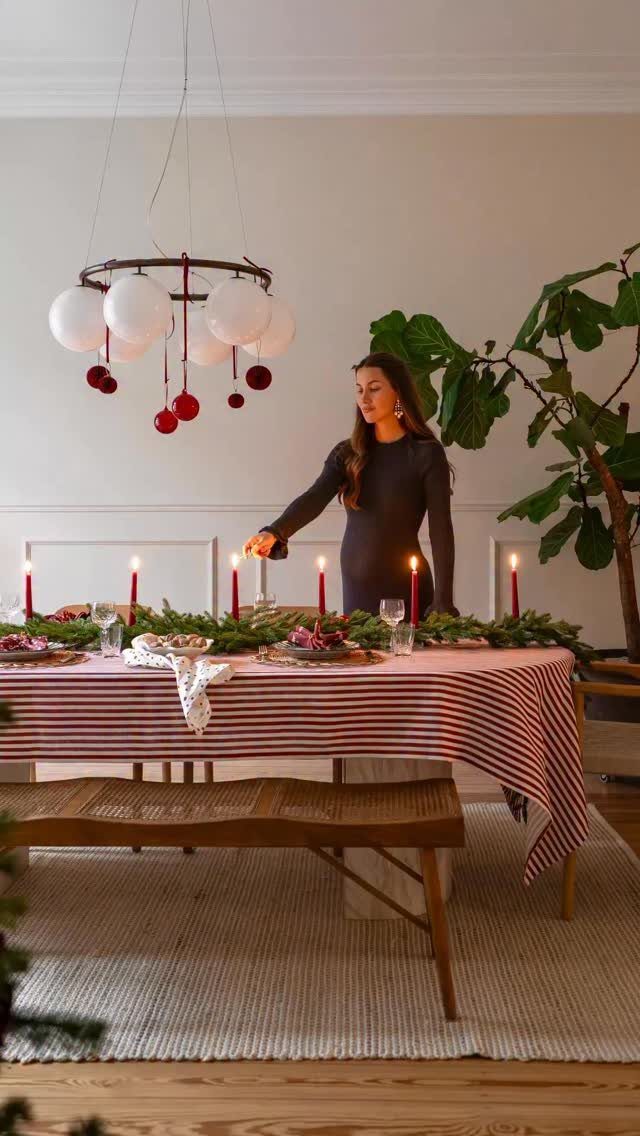 A woman sets a dining table decorated with a green garland and red candles.