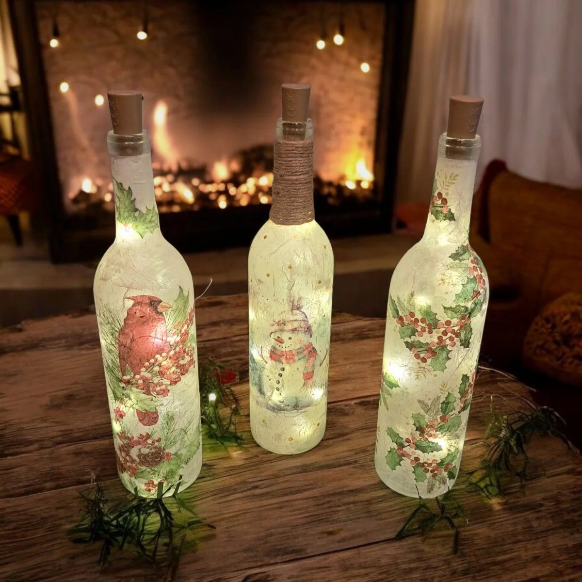 Three illuminated wine bottles decorated with festive decoupage designs sitting on a wooden table.