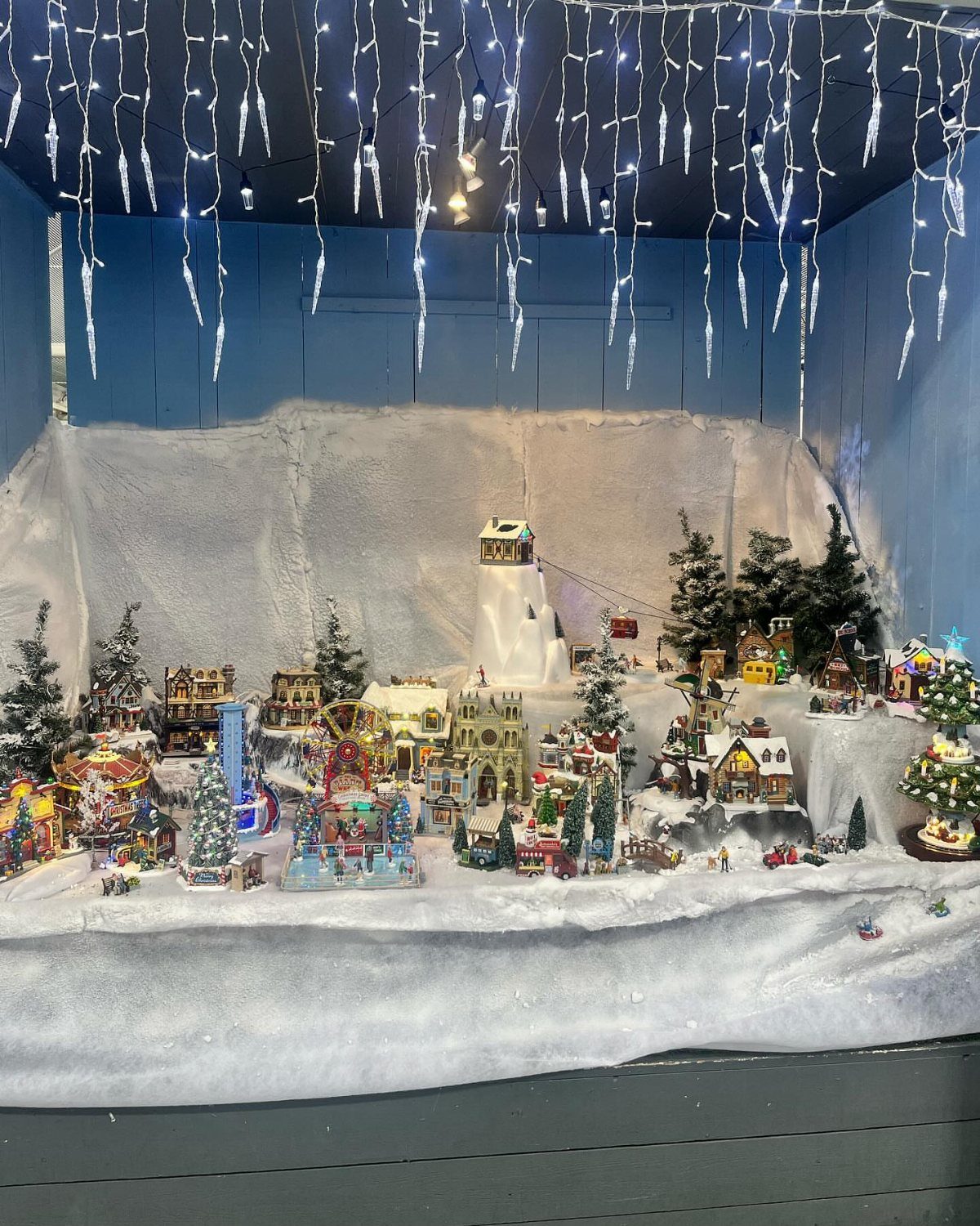 Miniature Christmas village with lighted houses and snowy landscape under hanging icicle lights.