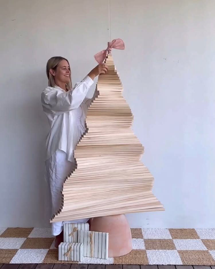 A woman standing beside a large, modern wooden Christmas tree made of stacked light wood slats.