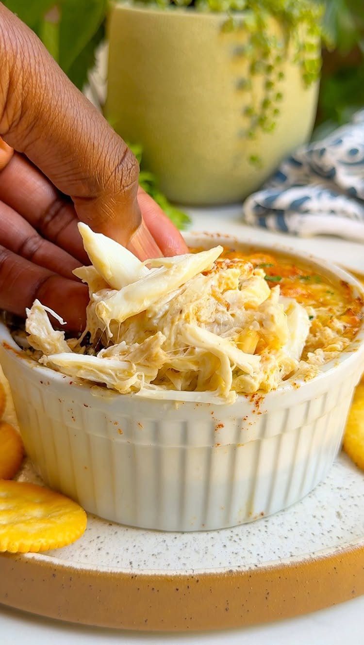 A hand dipping a cracker into a white ramekin of creamy hot crab dip.