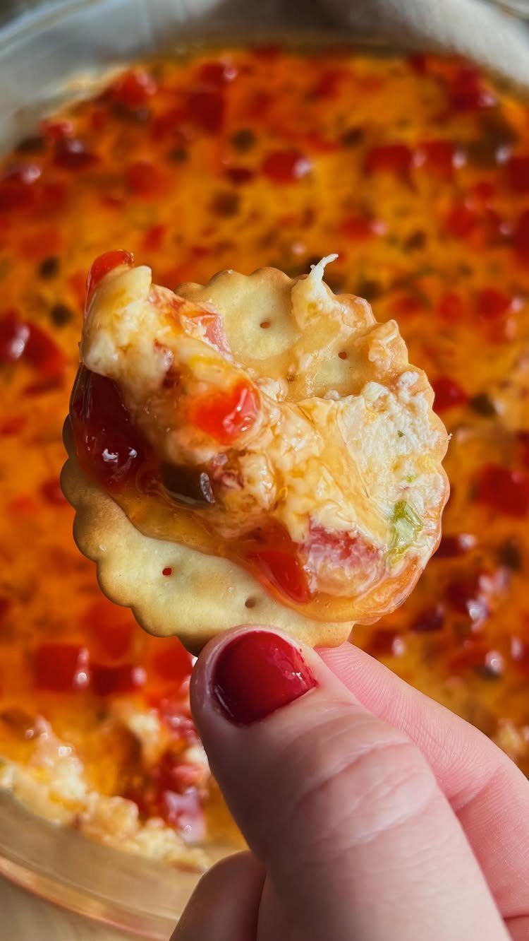 A hand holding a cracker topped with creamy pepper jelly cheese dip over a dish of more dip.