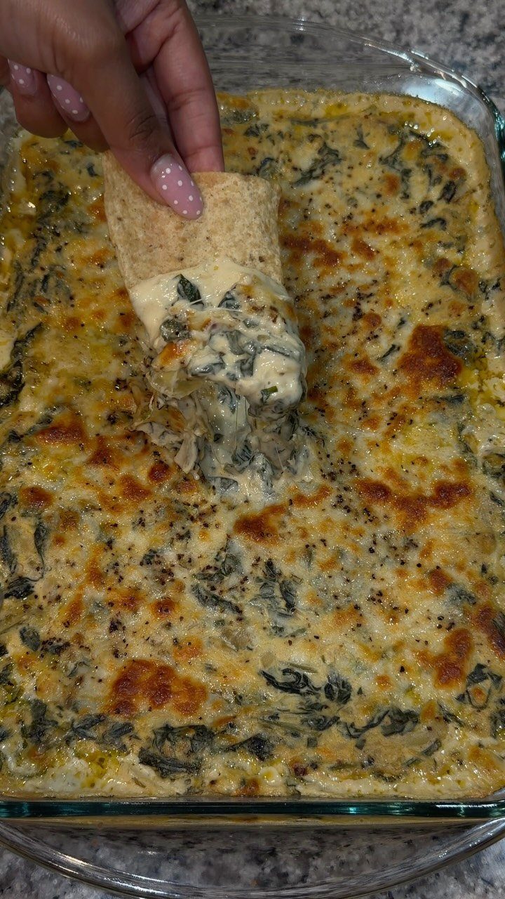 A hand dipping a tortilla chip into a warm, cheesy spinach artichoke dip.