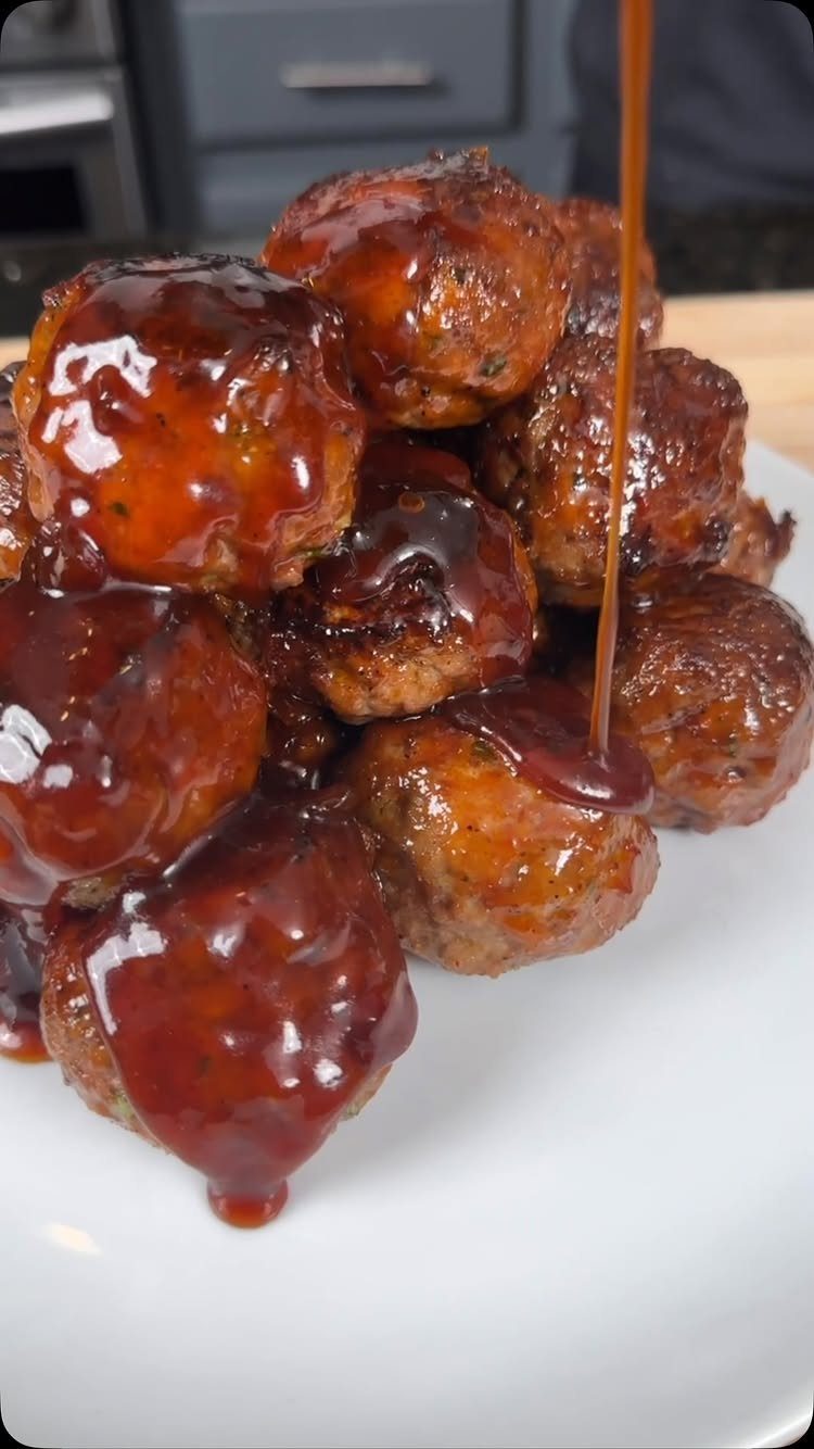 Glazed meatballs on a white plate with dark sauce being poured over the pile.