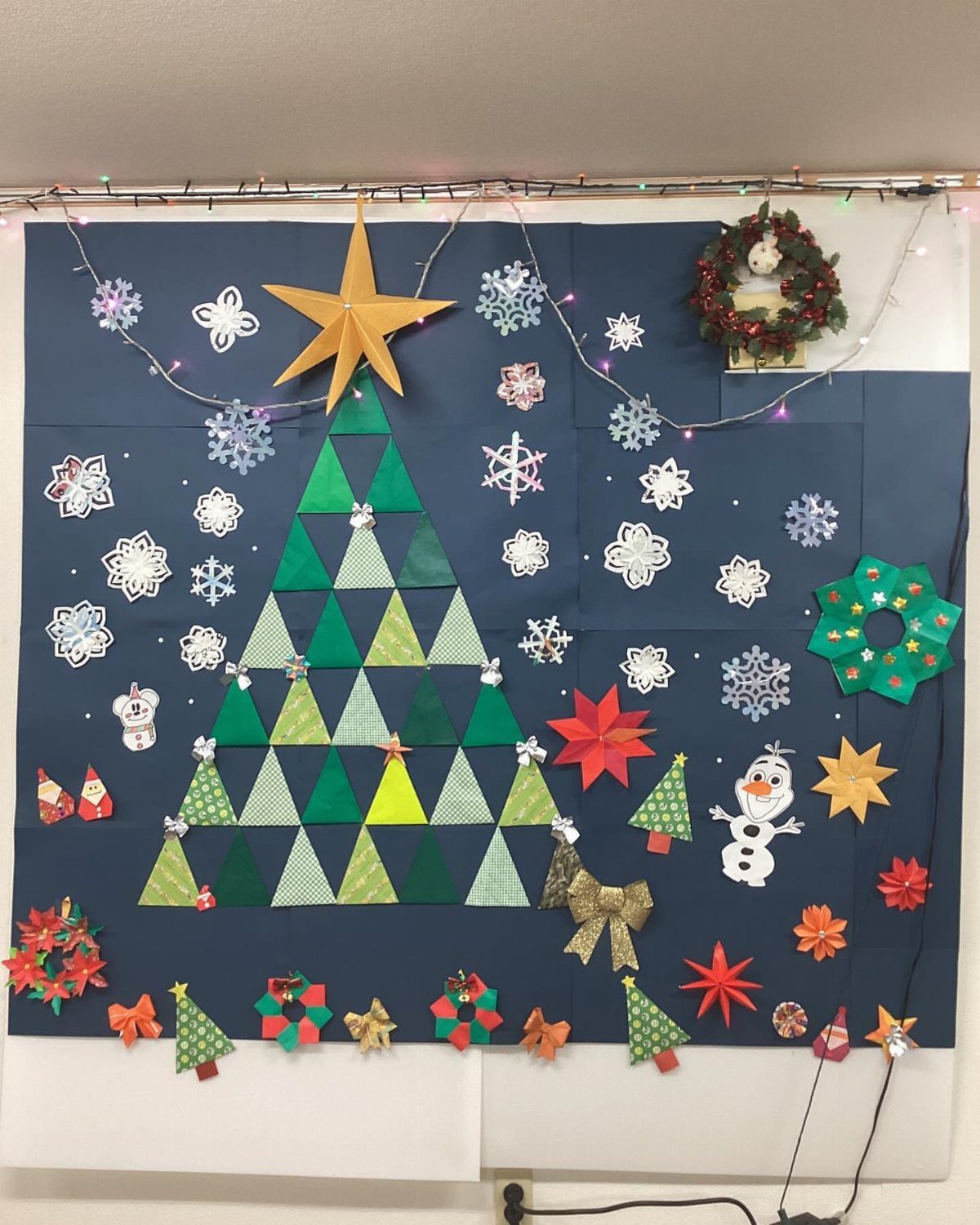 8 Christmas Tree Wall Ideas: String Lights to Pallet Builds 5 Paper Christmas tree made of green triangles on a blue wall with snowflakes and stars.