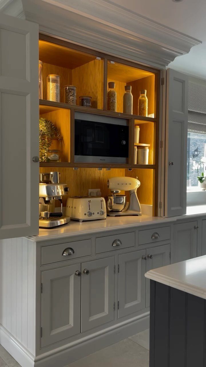 Open grey kitchen cabinets revealing wooden shelves with a microwave, toaster, and various kitchen appliances.