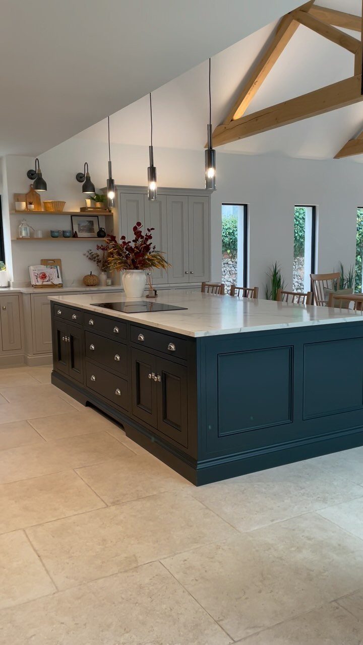 A dark blue kitchen island with a marble top stands in a bright room with timber beams.