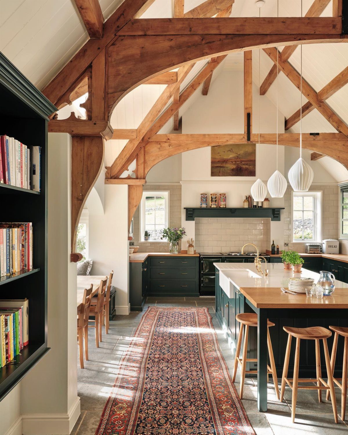 A rustic kitchen with large wooden ceiling beams, dark green cabinets, and a long patterned rug.