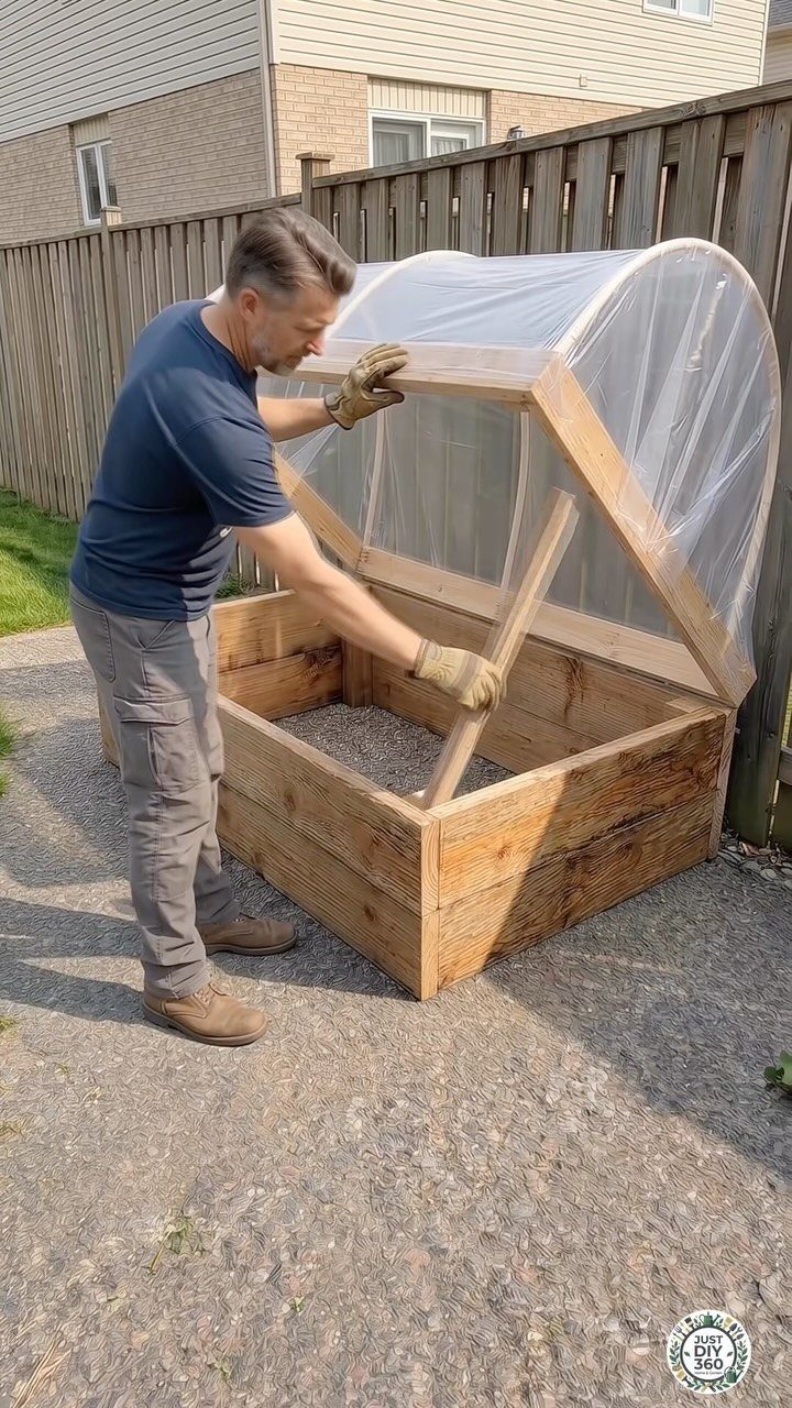 A man building a wooden raised garden bed with a clear plastic arched cover outdoors.