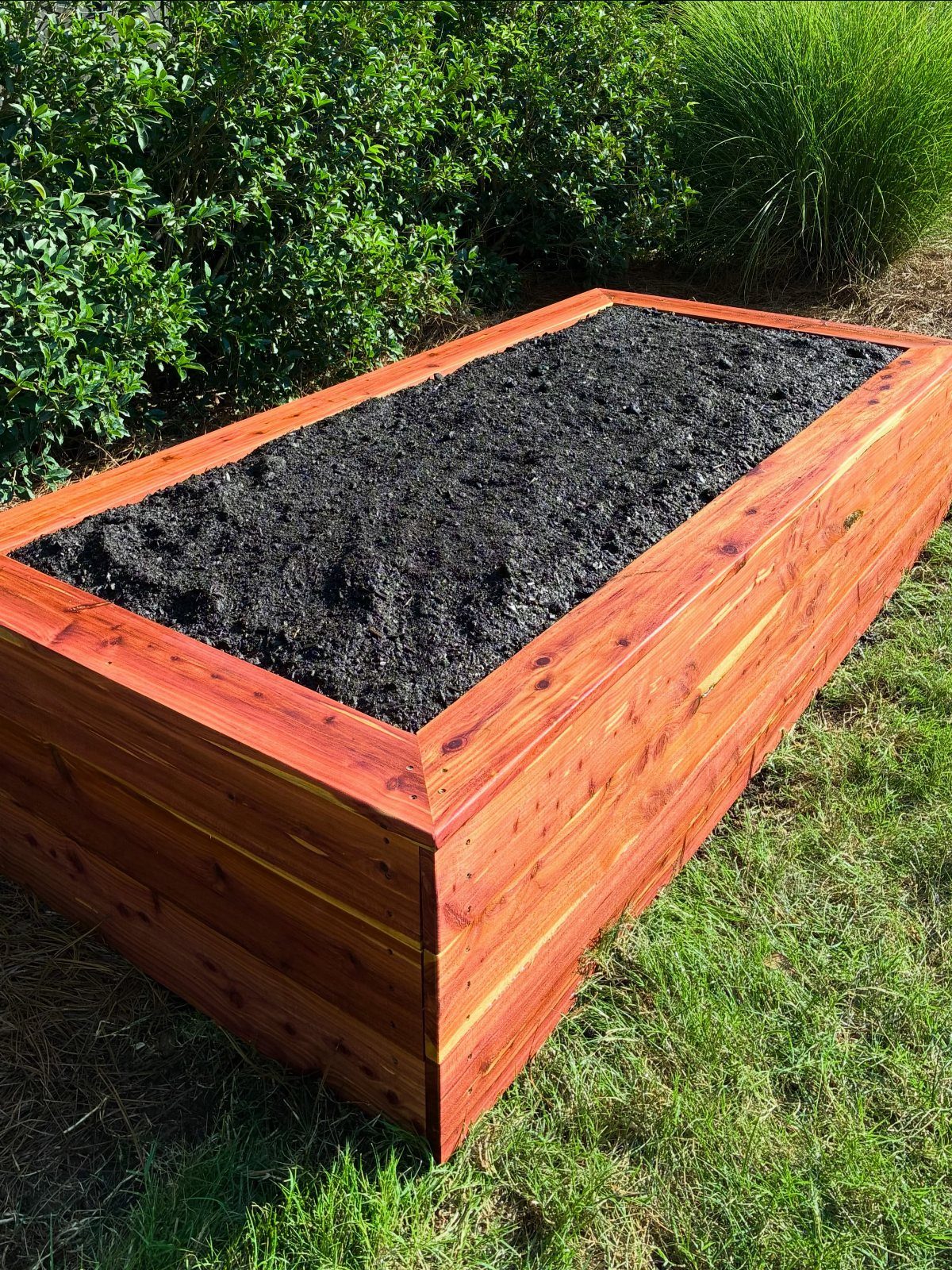 A rectangular cedar raised garden bed filled with dark soil sitting on a green lawn.