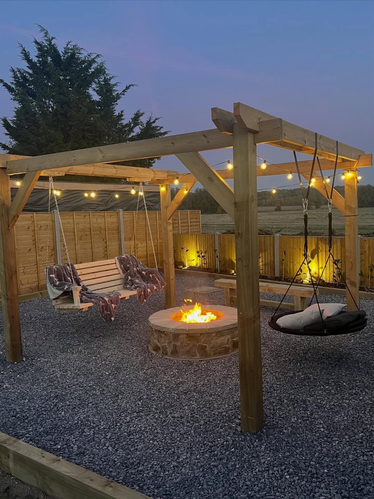 A stone fire pit glowing under a wooden pergola with hanging swings and string lights.