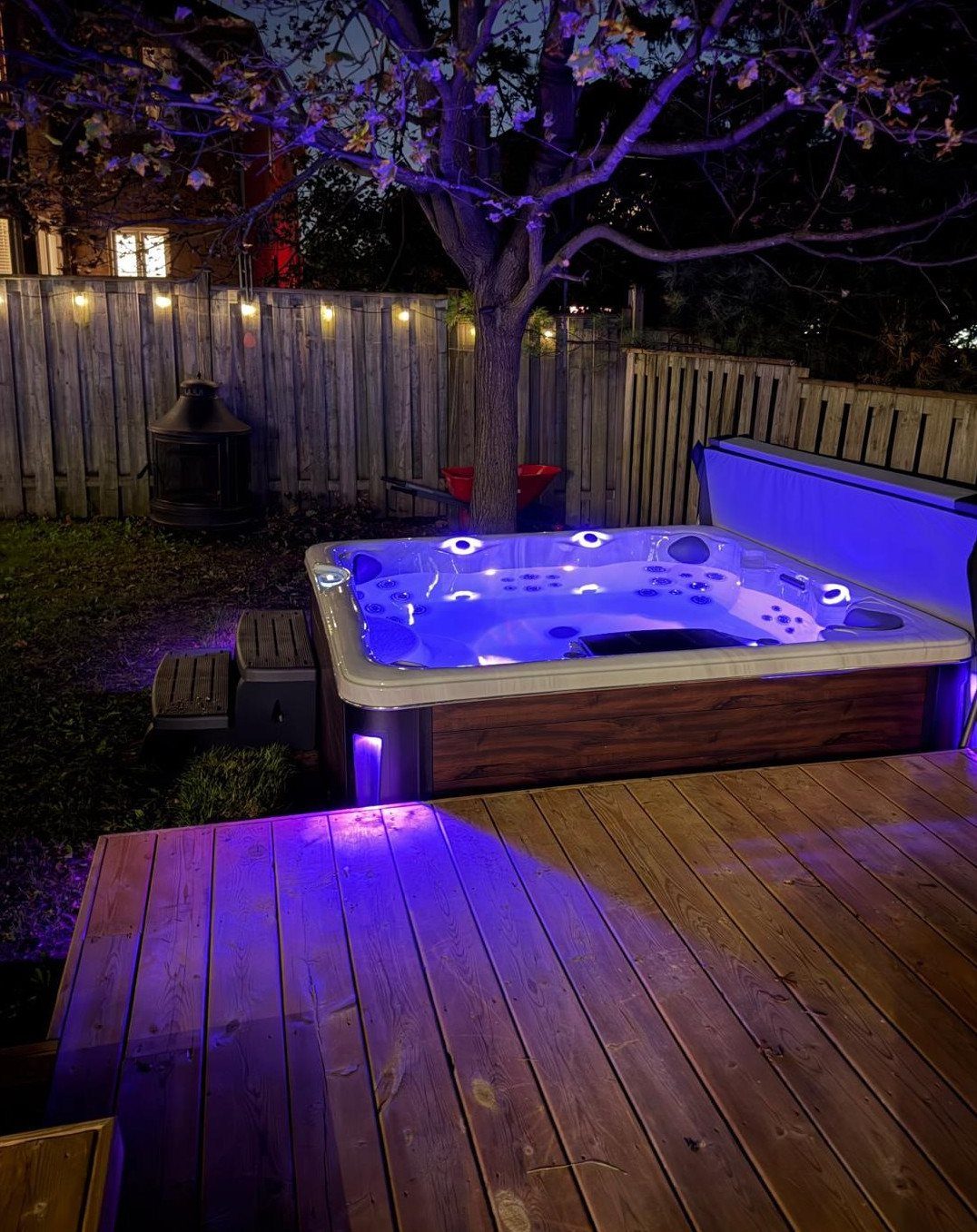 A glowing blue hot tub sits on a wooden deck in a dark backyard at night.
