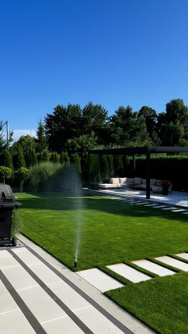 A green lawn with a water sprinkler in front of a black pergola and outdoor seating.