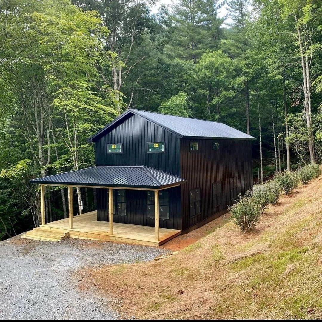 10 Ideas for Farmhouse Shed Home Style 6 A black two-story farmhouse shed with a wooden porch nestled in a lush green forest.