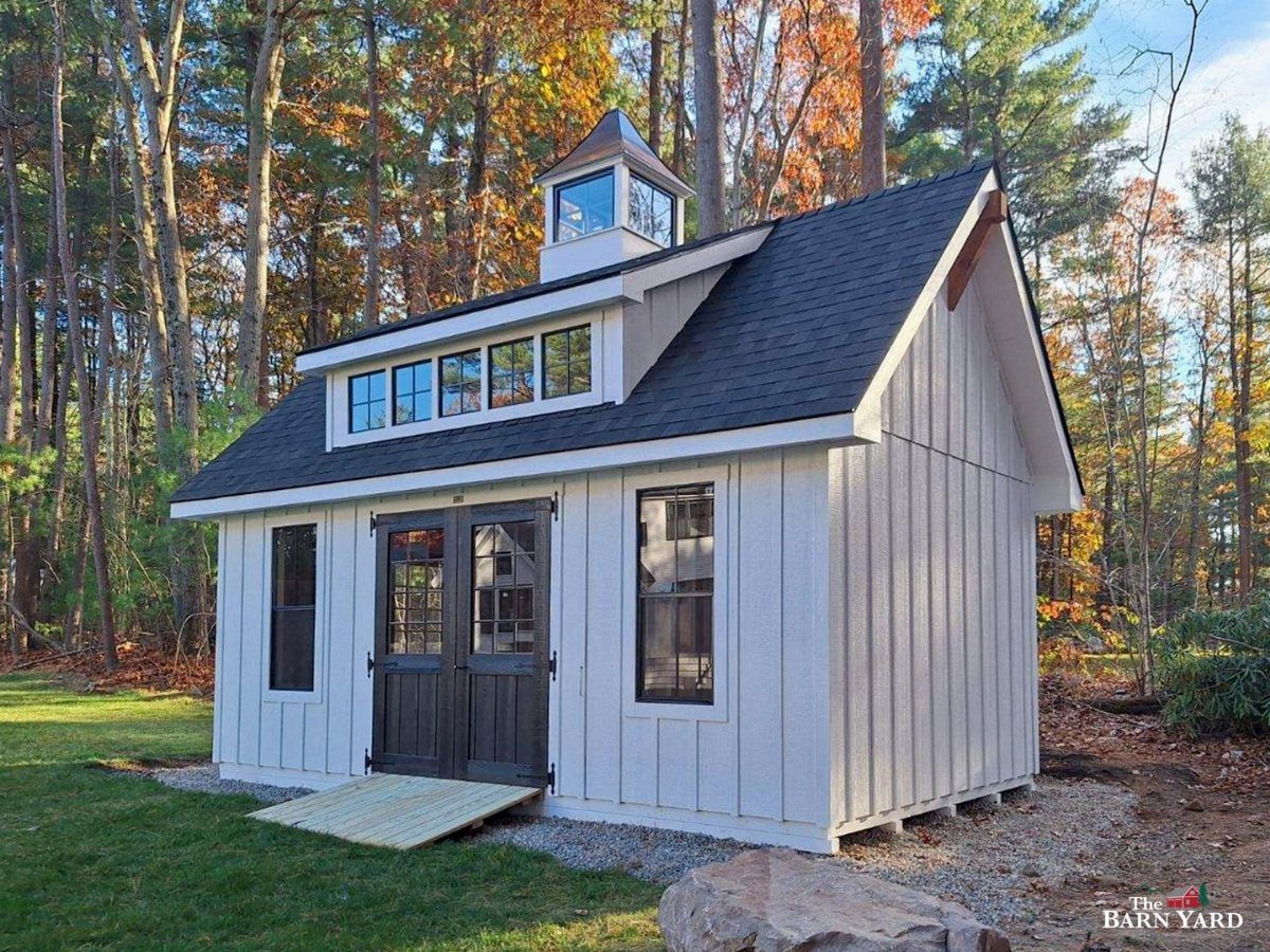 10 Ideas for Farmhouse Shed Home Style 1 White farmhouse shed with a large shed dormer and cupola in a forest setting.