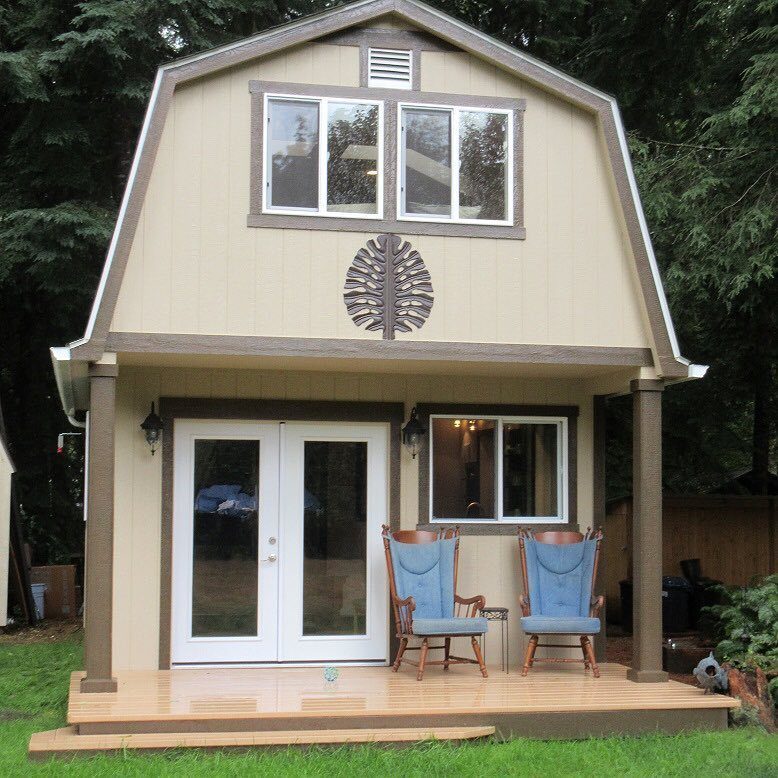 10 Ideas for Farmhouse Shed Home Style 7 A two-story tan farmhouse style storage shed with a small front porch and blue chairs.