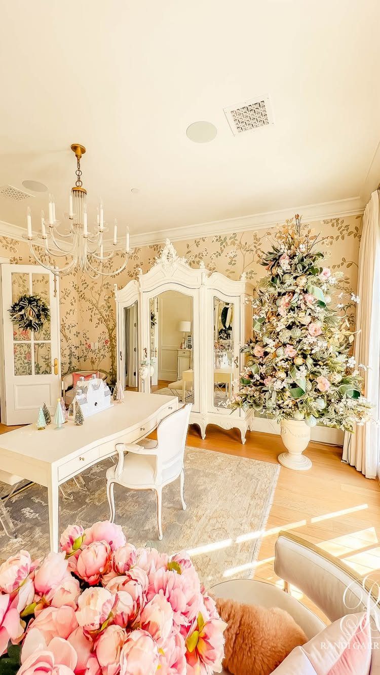 A large decorated Christmas tree stands in a bright room with floral wallpaper and white furniture.