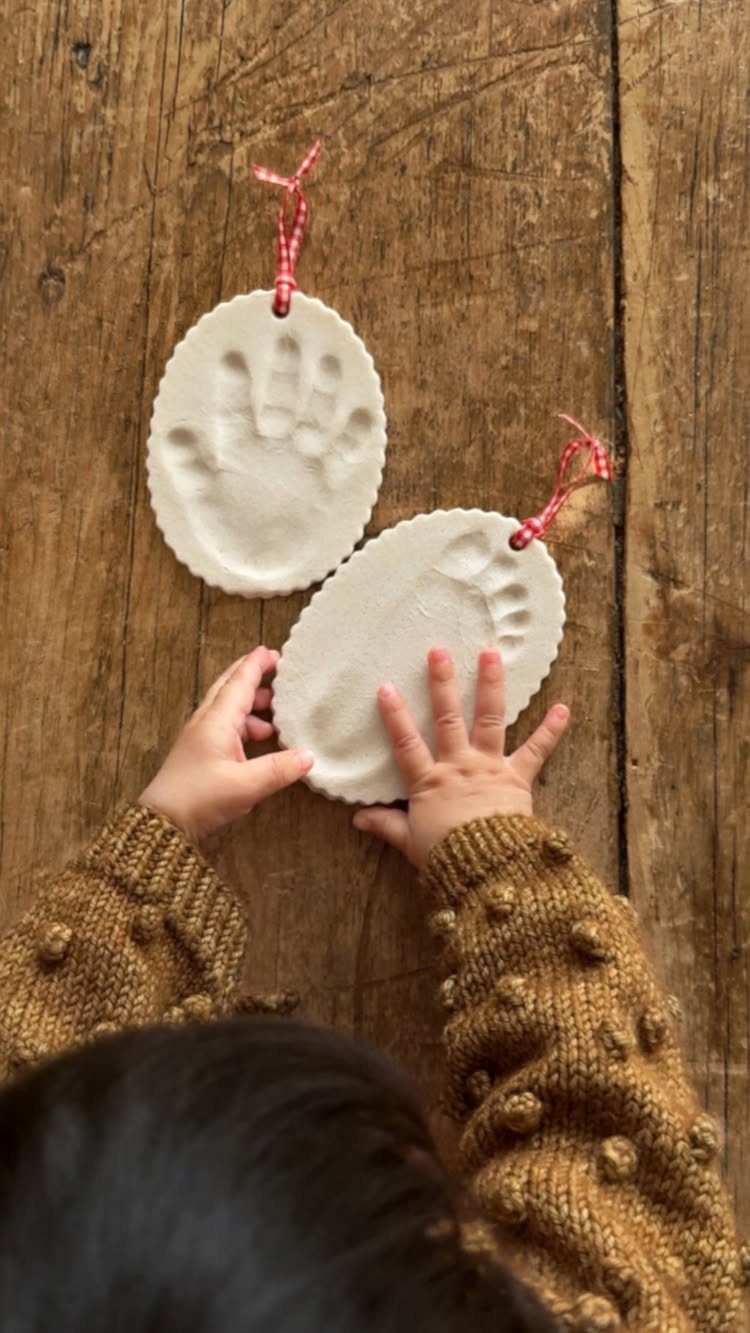 Child holding two white cornstarch dough ornaments with handprint impressions on a wooden table.