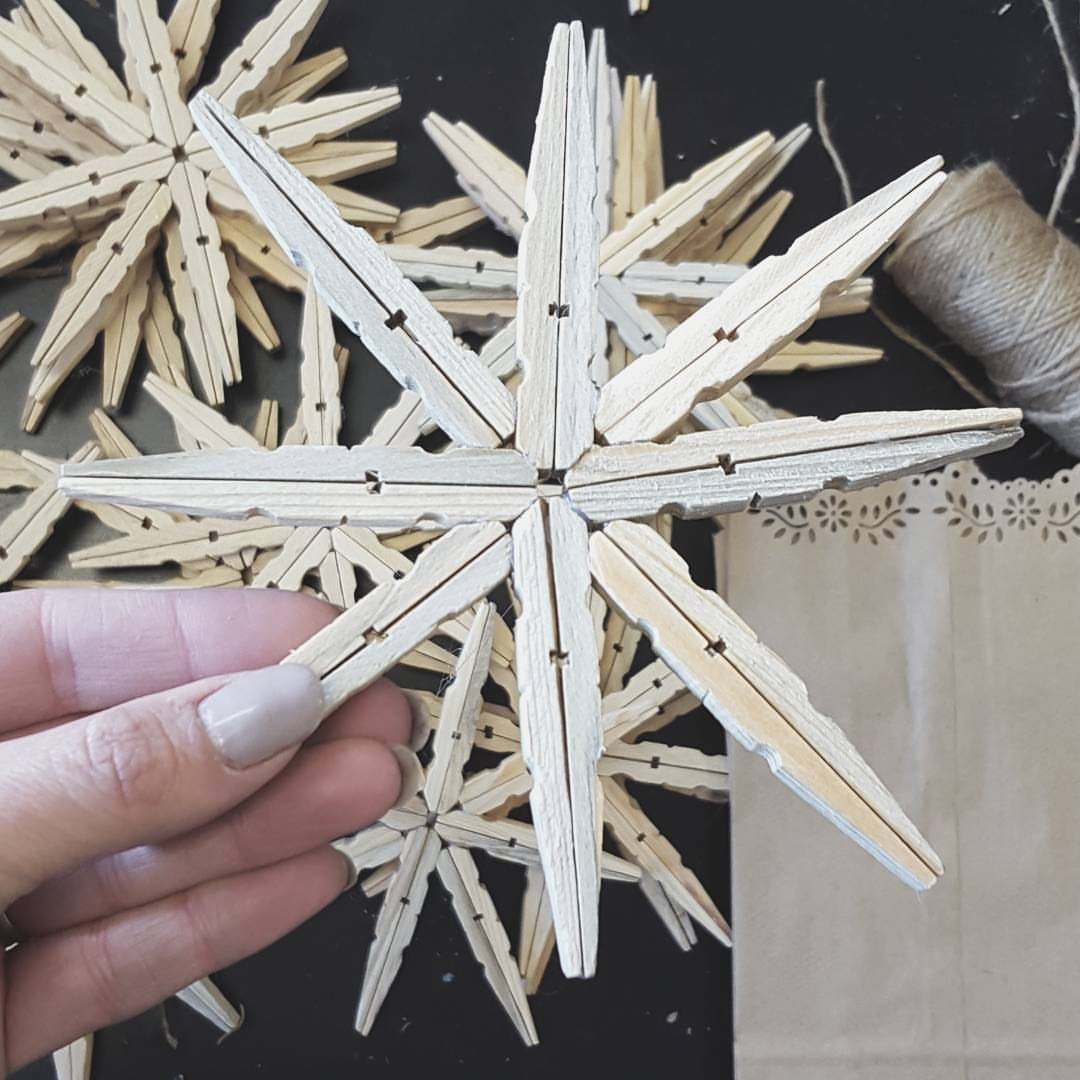 A hand holding a snowflake ornament crafted from multiple light wooden clothespins on a dark background.