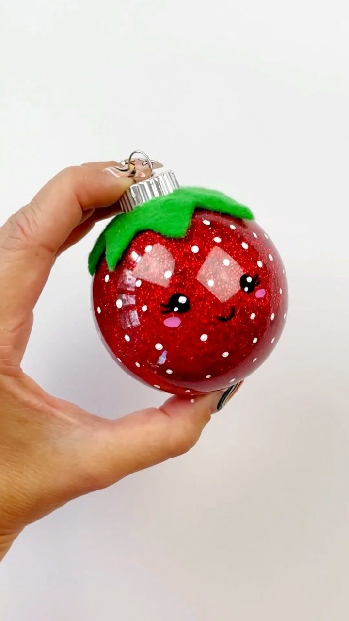 A hand holding a red glittery strawberry Christmas ornament with a cute smiling face.