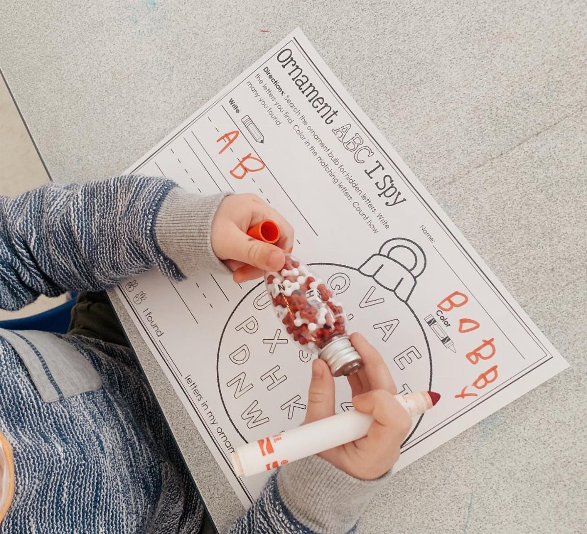 Child holding a clear plastic ornament filled with small red and white items over an activity sheet.