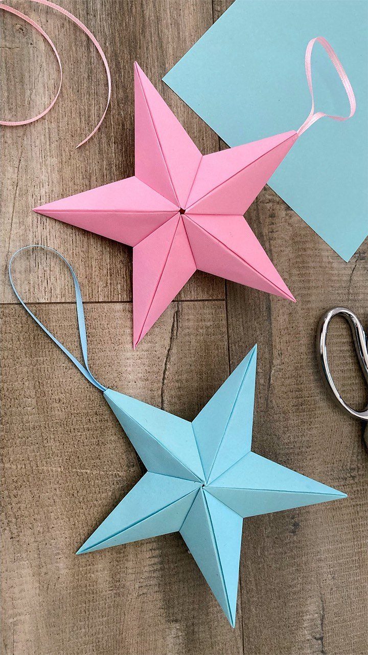 Pink and blue folded paper star ornaments resting on a wooden surface with craft supplies.