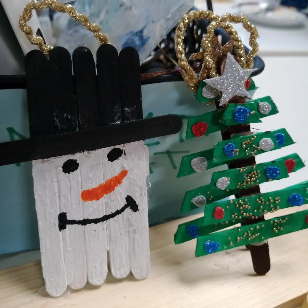 Handmade popsicle stick snowman and green sequined Christmas tree ornaments sitting on a table.