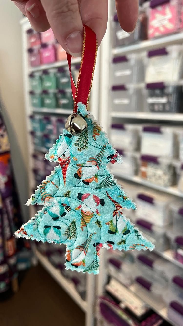 A hand holding a blue patterned fabric Christmas tree ornament with a red ribbon and bell.